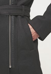 Mustat villcoat vyöllä ja hopealla vetskarilla, yksi käsi osittain sivutaskussa, näkyvää nahkareunusta vetskarin kohdalla.