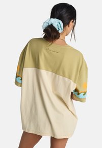 T-shirt surdimensionné en vert olive et crème, avec des motifs vifs de soleil et de vagues sur les manches. Tissu en coton doux avec une coupe décontractée.