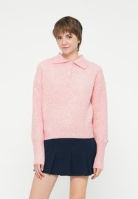 Pullover lavorato a maglia rosa chiaro con colletto, dotato di tre bottoni e maniche lunghe. Indossato sopra una gonna plissettata blu scuro.