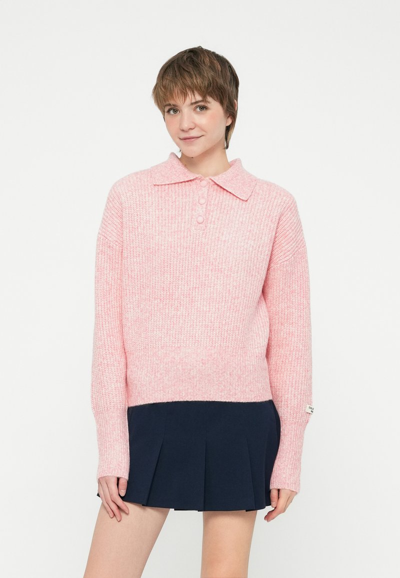 Pullover lavorato a maglia rosa chiaro con colletto, dotato di tre bottoni e maniche lunghe. Indossato sopra una gonna plissettata blu scuro.