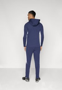 Marine trainingspak met capuchon, voorzien van witte zijkanten. Gemaakt van zachte stof, slim fit, met elastische manchetten en bijpassende joggers. Inclusief zwarte sportschoenen.