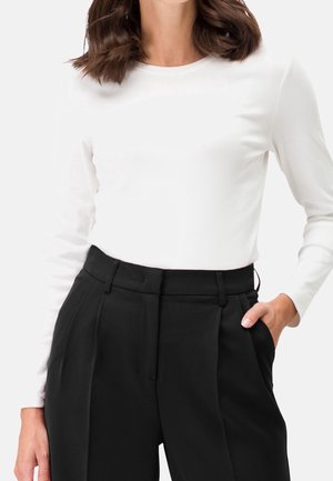 Femme portant une chemise blanche ajustée à manches longues rentrée dans un pantalon noir plissé taille haute, avec une main dans la poche.