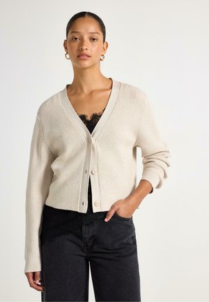Giovane donna con cardigan beige abbottonato sopra un top in pizzo nero e pantaloni scuri, in piedi davanti a uno sfondo chiaro e uniforme.