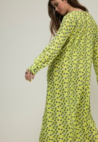 Langærmet, flydende kjole i lys gul med blomstermønster i grøn og hvid. Stoffet ser let ud; har knapmanchetter.