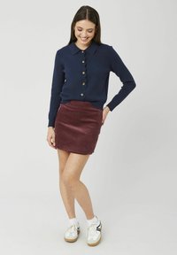 Jeune femme portant un cardigan bleu marine boutonné et une mini-jupe en velours côtelé bordeaux, debout, une main sur la hanche et souriante.