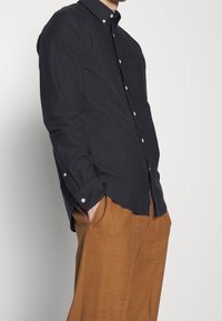 Camicia nera a maniche lunghe con bottoni in cotone. Presenta una tasca sul petto, bottoni bianchi e un orlo arcuato. Abbinata a pantaloni marroni texture.
