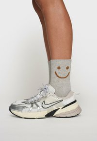 Srebrne i białe sneakersy z czarnymi akcentami, w połączeniu z jasnoszarymi skarpetkami z brązowym wzorem uśmiechniętej buzi.