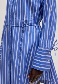 Blau gestreiftes Hemdkleid mit weißen vertikalen Streifen, tailliertem Gurt, geknöpften Manschetten und lockerer Passform. Hergestellt aus glattem Stoff.