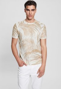 Šviesiai smėlio spalvos trumpomis rankovėmis marškinėliai su auksinės spalvos palmių lapų spauda. Minkšta tekstūra, apvalios iškirptės dizainas, derinami su baltais džinsais.