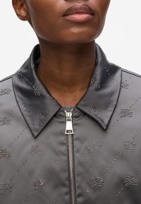 Giacca in satin grigio con chiusura a zip, rifinita con dettagli simili a perline e un colletto distintivo, caratterizzata da un motivo superficiale testurizzato.