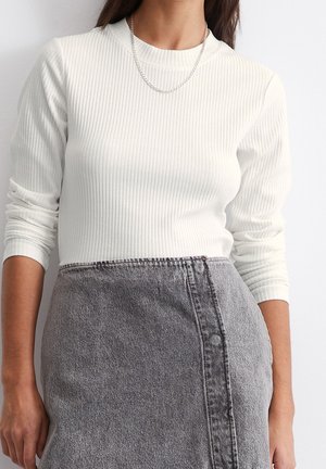 Frau trägt ein weißes, geripptes Langarmshirt, eine silberne Halskette mit Kette und einen hoch taillierten grauen Jeansrock mit sichtbaren Nahtdetails.