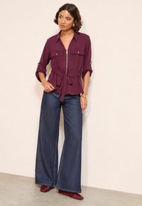 Veinihall peplum-bluus, millel on kahepoolne lukku ja rullitud varrukad, kahe rinnataskuga, kaetud laia jalgadega tumedate denimpükstega ja burgundia värvi kingadega.