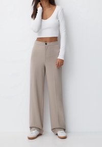 Biała crop top z długim rękawem i dekoltem w serek, w połączeniu z beżowymi spodniami typu wide-leg z przodem zapinanym na guzik i pionowymi szwami. Białe sneakersy.