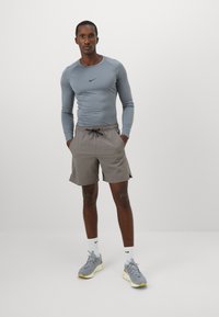 En atletisk mand står med hænderne i lommerne iført en tætsiddende langærmet grå Nike trøje, grå shorts, hvide Nike strømper og grå sneakers.