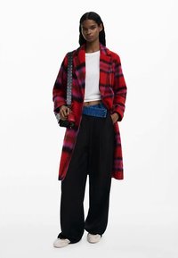 Piros és lila kockás, laza szabású kabát fehér felső és fekete, bőszárú nadrág fölött viselve. Díszített pántos táska és világos színű lábbeli kíséretében.