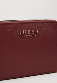 Burgunderfarbener strukturierter Reißverschluss-Wallet mit goldfarbenem „GUESS LOS ANGELES“-Logo auf der Vorderseite.