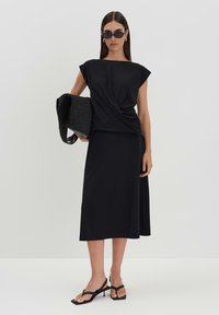 Haut drapé noir avec des manches courtes et une jupe mi-longue. Texture lisse, design minimaliste, associé à un sac noir texturé et des sandales à lanières.