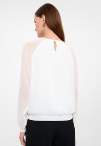 Witte blouse met lange, semi-doorzichtige mouwen, gathered taille en een sleutelgaatje aan de achterkant dat is vastgezet met een kleine knoop. Subtiel textuurpatroon.