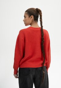 Rode geribbelde trui met een ronde halslijn, losse pasvorm en lange mouwen; gecombineerd met zwarte denim broek. Lange gevlochten haren vallen langs de rug.