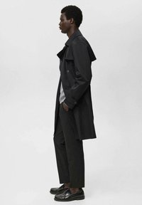 Abrigo negro tipo trench con detalle de capa, cierre de botón y bolsillos laterales. Llevado sobre un suéter gris y combinado con pantalones negros y mocasines.
