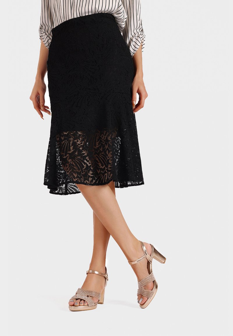 Gonna midi nera in pizzo con motivo floreale, caratterizzata da un orlo a scallop; abbinata a sandali con tacco alto metallici dorati e con cinturini.