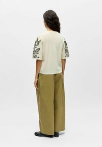 Femme aux cheveux longs et foncés portant un haut crème à manches courtes avec une broderie noire sur les manches, un pantalon large olive et des chaussures noires, vue de dos.