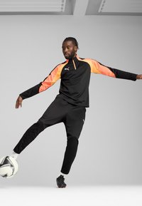 Fato de treino atlético preto com detalhes em laranja, mangas longas e colarinho alto. Modelo executa um chute com uma bola de futebol branca.