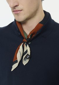 Polo en maille bleu marine avec une écharpe à motifs brun, vert et crème nouée au cou. L'écharpe présente des rayures et une petite étiquette avec un logo.
