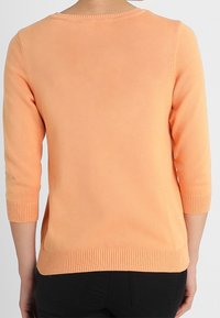 Pull de couleur pêche avec des manches trois-quarts, ourlet et encolure côtelés, présentant une texture lisse et une coupe légèrement ajustée.