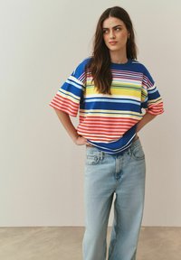 Ženska z dolgimi rjavimi lasmi, oblečena v pisano široko progasto majico in svetlo modre kavbojke, stoji z rokami na bokih pred enobarvnim ozadjem.