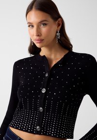 Czarny krótki kardigan o prążkowanej fakturze, ozdobiony srebrnymi ćwiekami, które przechodzą od gęsto rozmieszczonych do rzadszych. Posiada pięć srebrnych guzików.