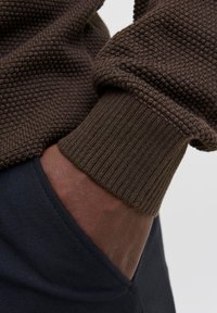 Temna koža roka vtaknjena v žep mornarsko modrih hlač, oblečena v rjave teksturirane pletene pulover s rebrastimi manšeti.