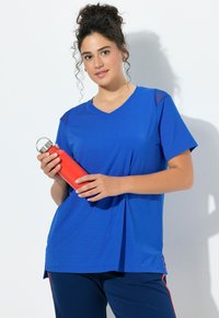 Mladá žena v modrém sportovním oblečení drží červenou kovovou láhev na vodu, stojící na bílém pozadí.