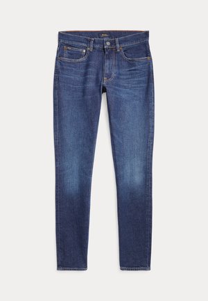 Tumši zili pieguļoši džinsi ar klasisku piecu kabatu dizainu un pogas aizdari, noklāti uz baltas virsmas.