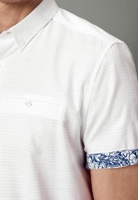 Camicia bianca a maniche corte con un tessuto a trama, dotata di taschino sul petto e motivo floreale blu sui polsini.