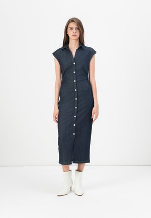 Robe en denim avec des manches courtes, fermeture à boutons sur le devant, longueur au genou, de couleur bleu foncé, associée à des bottines blanches. Texture lisse.