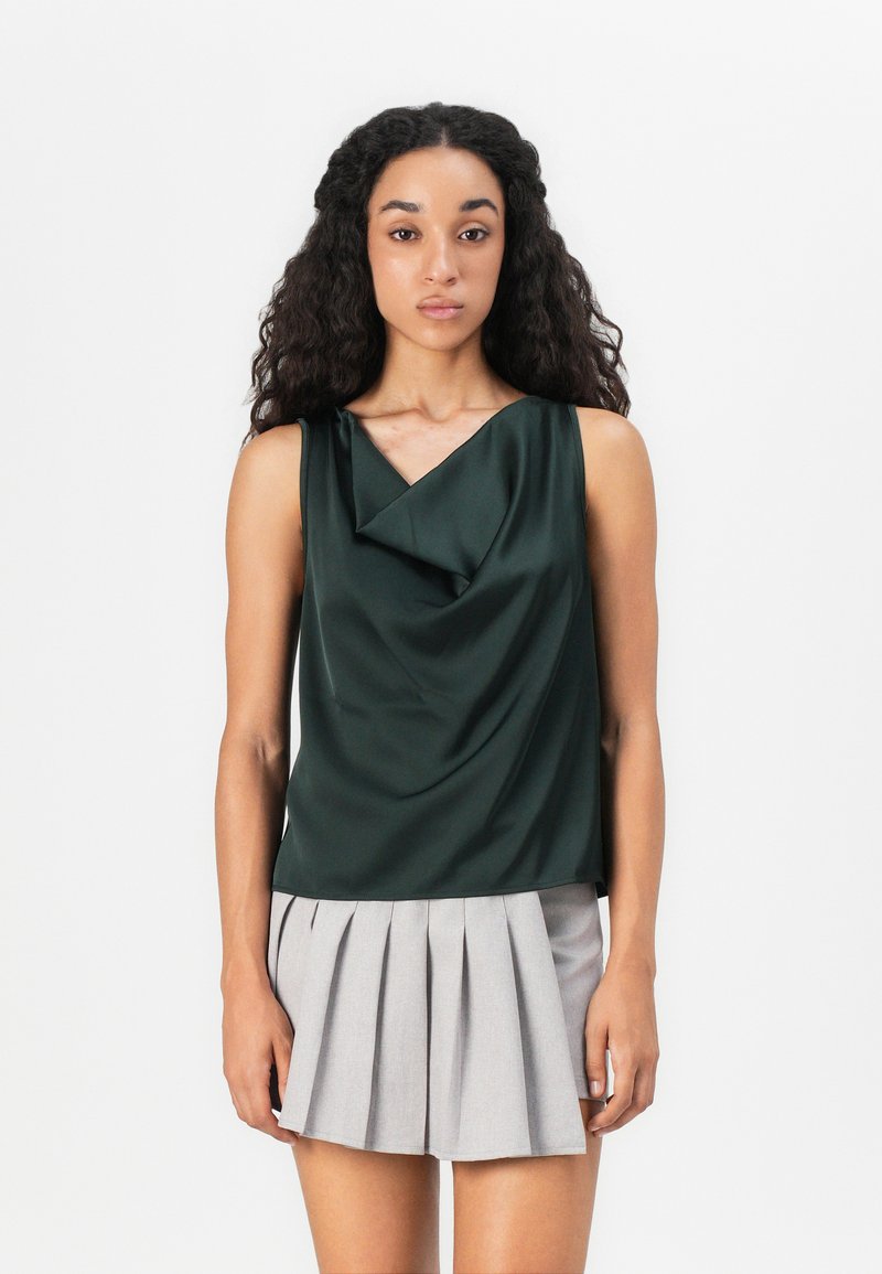 Top sin mangas de color verde oscuro en tejido suave con escote drapeado, combinado con una falda corta plisada de color gris claro.