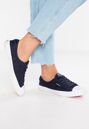 Sneakers in tela blu navy con punte e suole in gomma bianca, dotati di lacci e di una patch con il logo sulla linguetta. Indossati con jeans chiari a vita corta.
