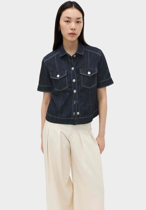 Vrouw met lang donker haar die een donkerblauw denim overhemd met korte mouwen en een keuze wijde, high-waisted crèmekleurige broek draagt.