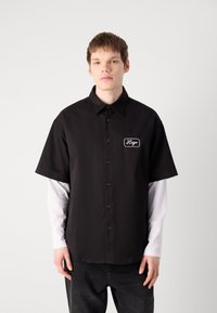 Zwarte korte mouwen shirt met een knoopsluiting aan de voorkant, voorzien van een witte geborduurde logo patch. Gelaagd over een wit langemouwen shirt.