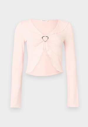 Ανοιχτό ροζ crop top με μακριά μανίκια, με κυματιστές άκρες και μεταλλικό δαχτυλίδι σε σχήμα καρδιάς στο κέντρο μπροστά.