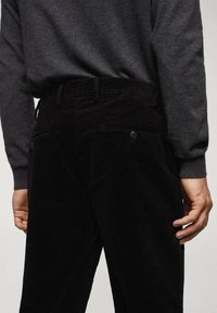 Personne portant un pantalon en velours côtelé noir avec des poches arrière boutonnées et une chemise à manches longues gris foncé, debout les mains détendues le long du corps.