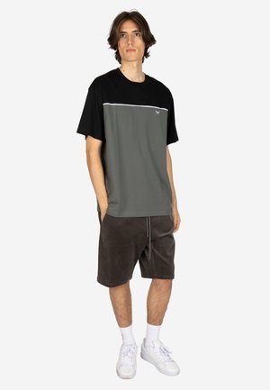 Young man stands with hands in pockets wearing black and gray short-sleeve shirt, dark shorts, white socks, and white sneakers on white background.