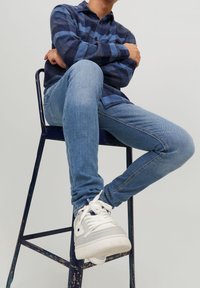 Camisa de franela a cuadros azul y azul marino, jeans ajustados azul claro, zapatillas blancas con detalles grises, sentado en un taburete de metal contra un fondo liso.