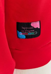 Czerwona bluza z ribbowanym dołem, z czarną naszywką. Naszywka ma kolorowe serca oraz biały napis "happiness in progress".