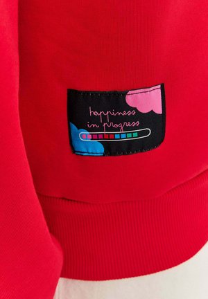 Czerwona bluza z ribbowanym dołem, z czarną naszywką. Naszywka ma kolorowe serca oraz biały napis "happiness in progress".