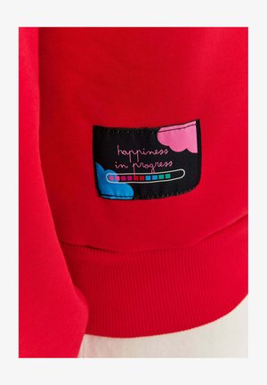 Czerwona bluza z ribbowanym dołem, z czarną naszywką. Naszywka ma kolorowe serca oraz biały napis "happiness in progress".
