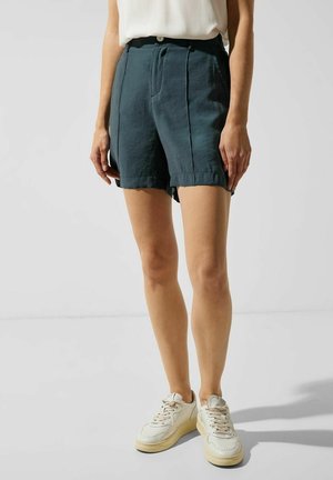 Vrouw met donkerblauwe korte broek, wit topje en witte sneakers, staand tegen een effen lichte achtergrond.