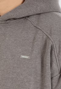 Grå hættetrøje lavet af blødt stof, med et metal logo mærke foran, hætte med snøre og raglanærmer.