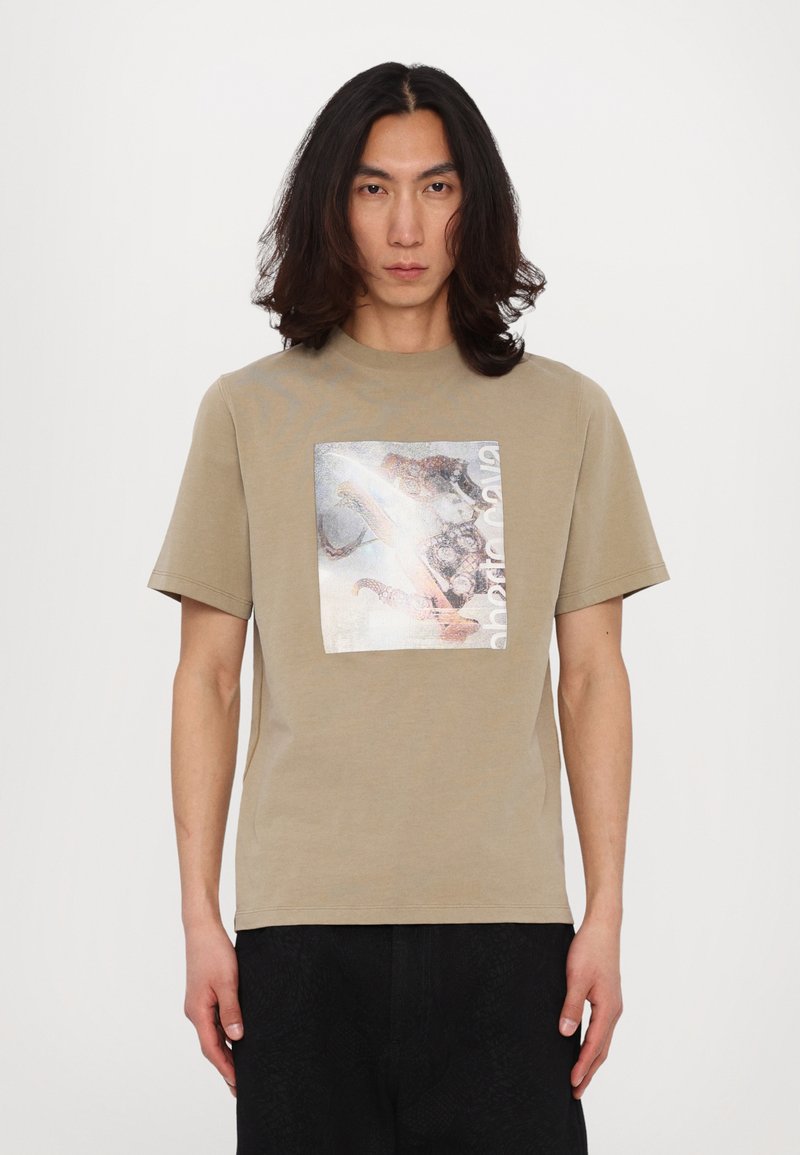 Camiseta de algodón beige con un estampado gráfico metálico cuadrado que presenta un diseño mixto. Mangas cortas y escote redondo, corte relajado.